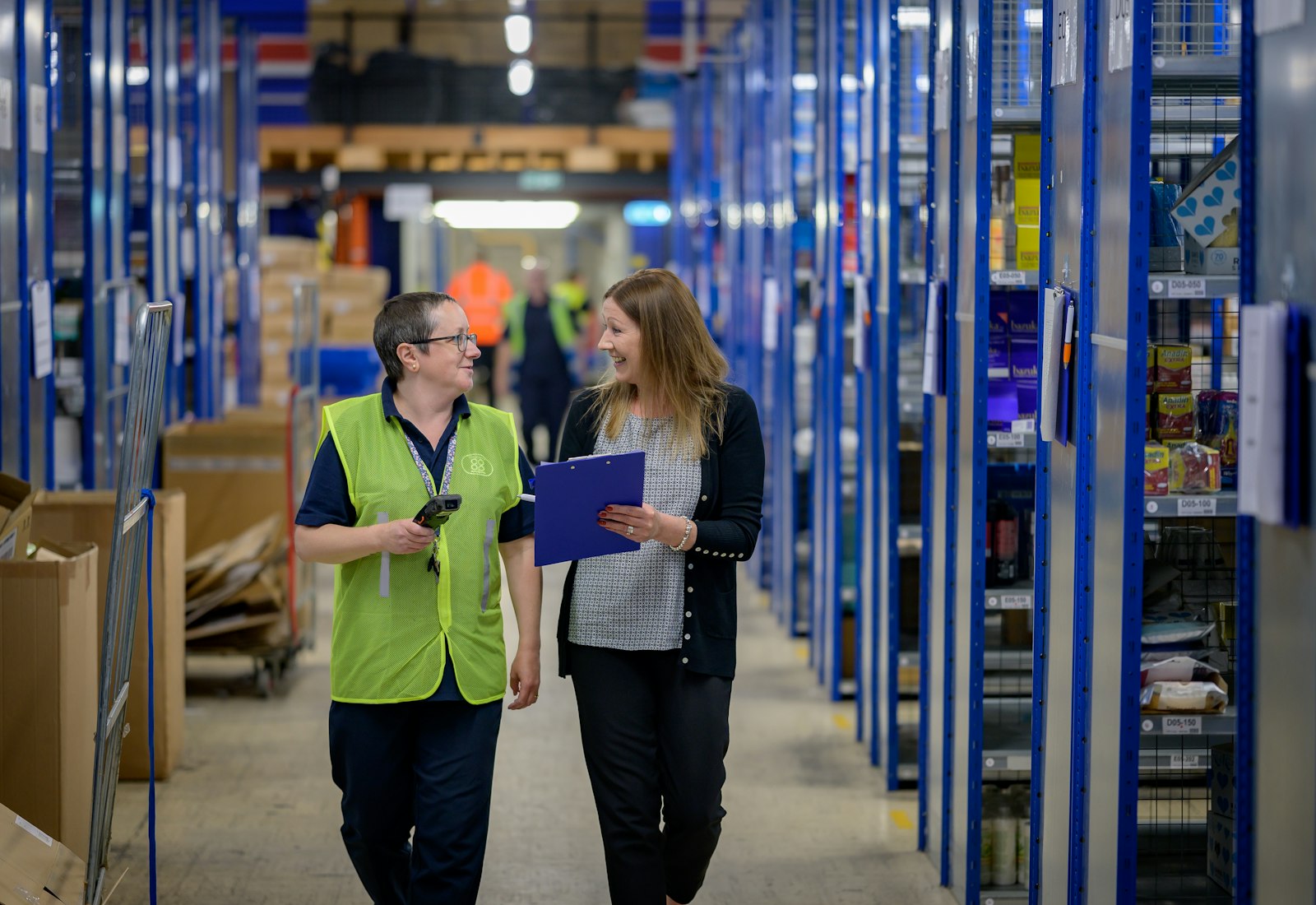 Salesforce Consumer Goods Cloud mit Beyond Digital: Zwei Mitarbeiterinnen mit Klemmbrett und Scanner gehen durch einen Lagergang mit blauen Regalen